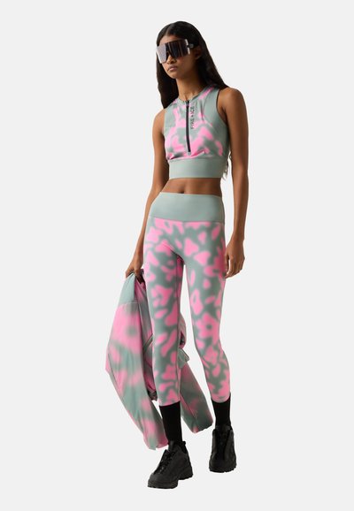 Frau trägt ein passendes pink-grau gemustertes Activewear-Set mit Crop-Top und Leggings, hält eine Jacke, trägt große schwarze Sonnenbrillen und schwarze Sneakers.