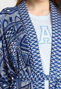 Niebiesko-biała szata z geometrycznymi wzorami noszona na jasnej koszuli z dużą literą "A" wyszytą niebieską nitką.