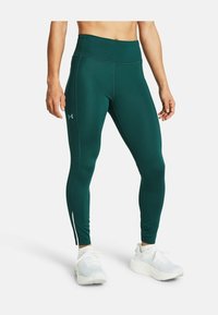 Osoba atletyczna w ciemnozielonych legginsach z wysokim stanem i białych sneakersach, stojąca na białym tle.