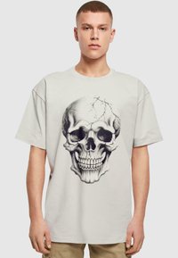 Lys grå kortærmet t-shirt med et stort sort-hvidt skull grafik i et pixeleret design med revnede detaljer.