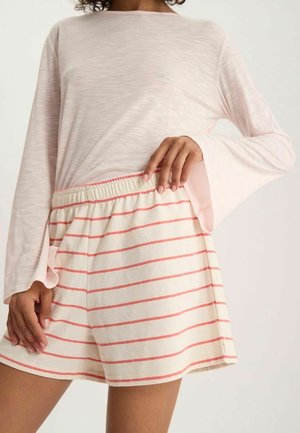 Femme portant un haut à manches longues rose clair et un short crème avec des rayures horizontales roses, debout avec la main sur la hanche.