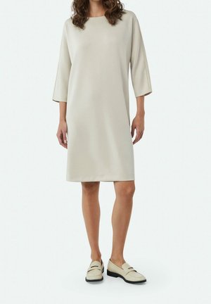 Kvinde iført en knælang, cremefarvet kjole med trekvartlange ærmer og matchende cremefarvede loafers mod en ensfarvet baggrund.