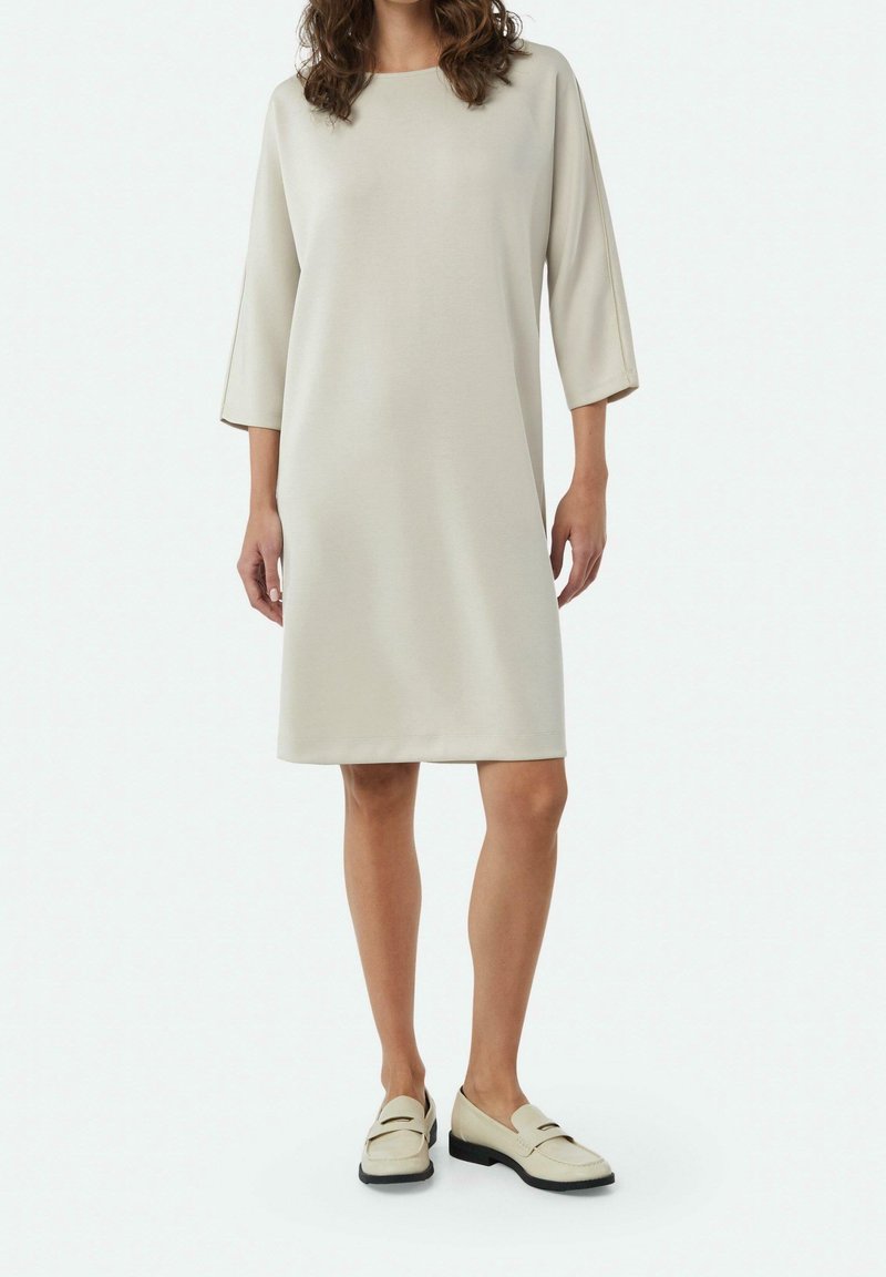 Femme portant une robe crème mi-longue à manches trois-quarts et des mocassins crème assortis sur un fond uni.