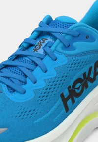 Sapatilha de corrida Hoka azul com parte superior em malha, atacadores azuis a condizer, logótipo preto de lado e sola branca com detalhe amarelo.