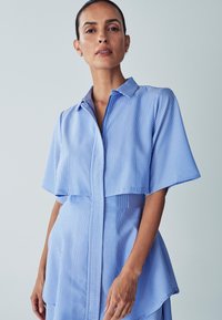 Blau-weiß gestreiftes Hemdblusenkleid mit Knopfleiste, kurzen Ärmeln und asymmetrischem Saum. Glattes Material mit leichtem Glanz.