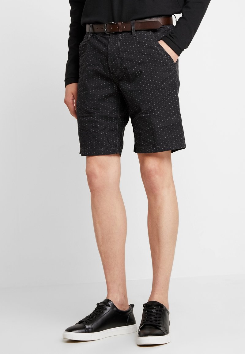 Pantalones cortos negros con un patrón de puntos, fabricados en tejido ligero, con bolsillos laterales y un dobladillo recto. Llevados con un cinturón marrón y zapatillas negras.