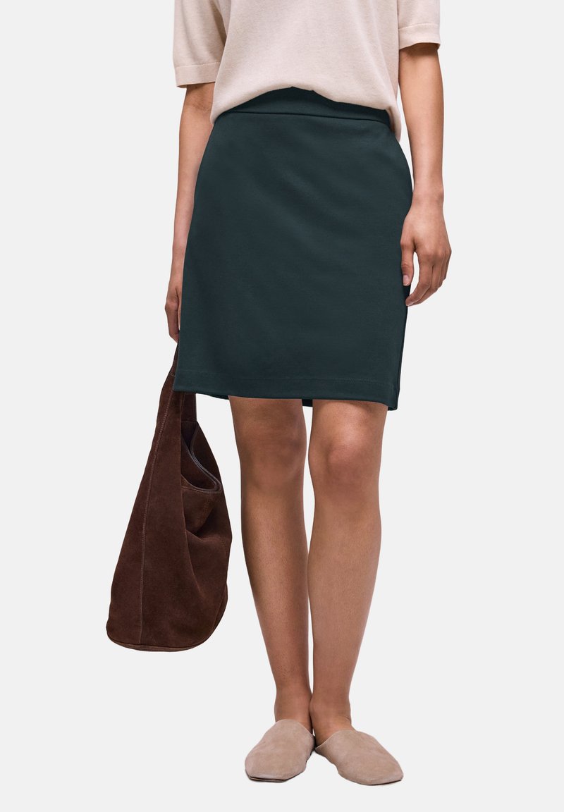 Femme portant une jupe vert foncé longueur genou, un haut beige, des chaussures beiges à enfiler, et tenant un grand sac cabas en daim marron.