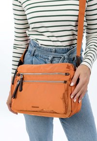 Person wearing blue jeans and a striped long-sleeve shirt holding an orange crossbody bag with multiple zipper pockets.