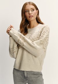 Krémový pletený sveter s textúrovaným vzorom, ktorý obsahuje geometrické akcenty, rebrované manžety a uvoľnený strih.