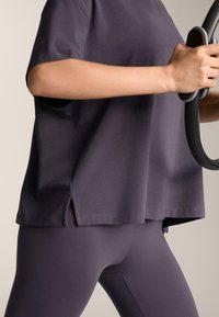 Osoba v tmavo fialovom športovom oblečení drží čierny Pilates kruh na úrovni pásu na jednofarebnom pozadí.