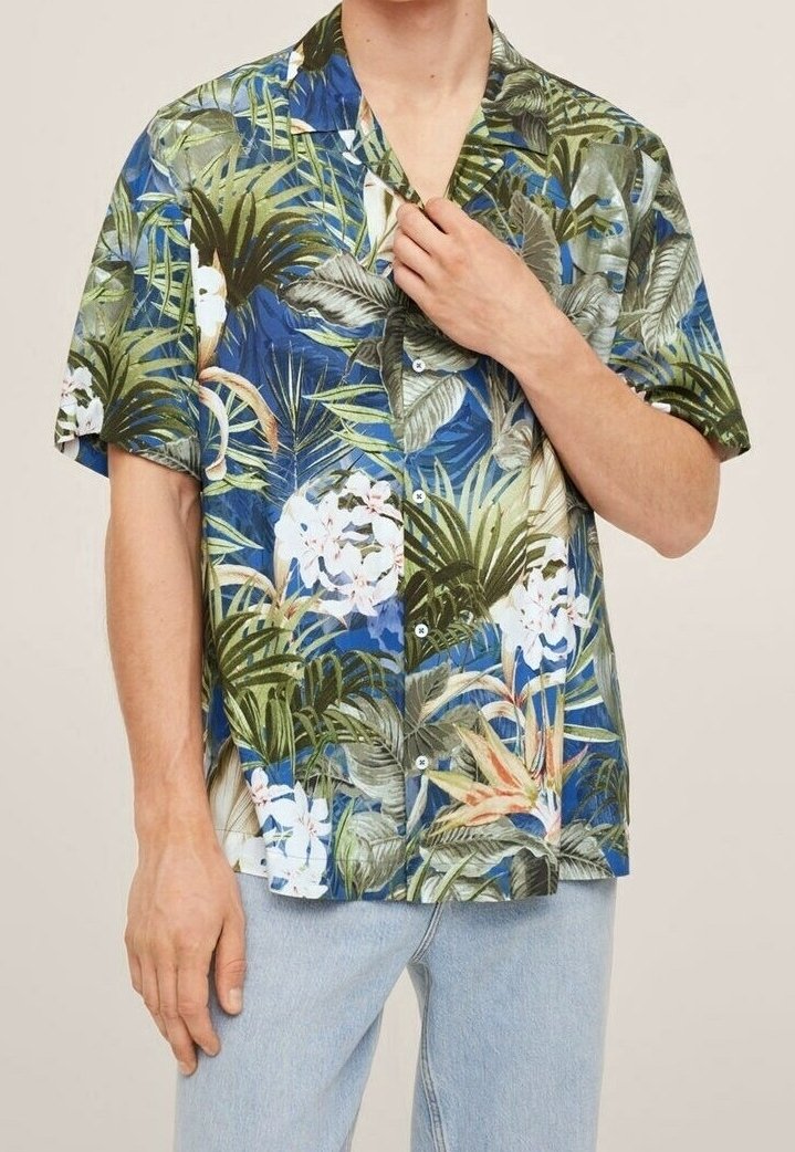 Homme portant une chemise à manches courtes boutonnée avec un motif floral tropical bleu, vert et blanc, associée à un jean bleu clair.