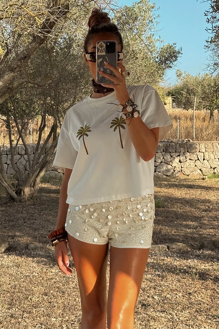 T-shirt blanc court avec des motifs de palmier, associé à un short crème scintillant orné de sequins, vu dans un cadre naturel en extérieur.