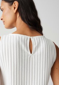 Top bianco texturizzato con righe verticali, scollatura ampia e chiusura a chiave sul retro con un piccolo bottone decorativo.