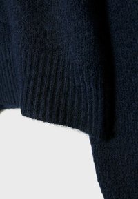 Maglione blu navy lavorato a maglia con superficie testurizzata e polsini a costine; il tessuto appare morbido con una leggera lucentezza su uno sfondo chiaro.