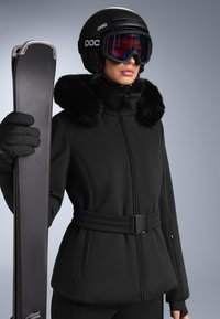 Crni skijaški jakna s krznom oko vrata, kroj prilagođen tijelu, pojas na struku. Skijaške naočale s višebojnim lećama, drži crne skije s teksturiranom površinom.
