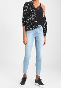 Blouse noire à pois, superposée à un caraco en dentelle noire, associée à un jean skinny bleu clair et des baskets noires. Textures lisses et design décontracté.
