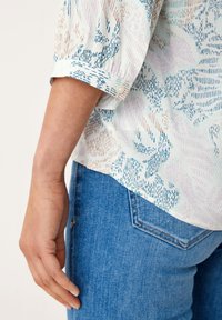 Blouse légère à manches courtes avec un motif abstrait multicolore dans des tons pastel. Dotée d'un poignet boutonné et d'un tissu fluide, portée avec un jean bleu.