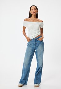 Valkoinen ribattu olkaimeton paita pitsireunalla; löysät siniset farkut korkealla vyötäröllä. Malli poseeraa kädet taskuissa ja käyttää tennareita.