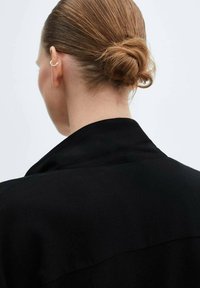 Giacca nera con colletto, vista da dietro. I capelli sono raccolti in uno chignon ordinato, con un orecchino a cuffia argentato visibile.