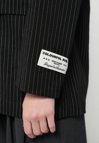Čierny sako s tenkými pásikmi, s bielou textilnou štítkou "COLOURFUL REEL" na manžete. Má štruktúrovaný dizajn a prispôsobený strih.