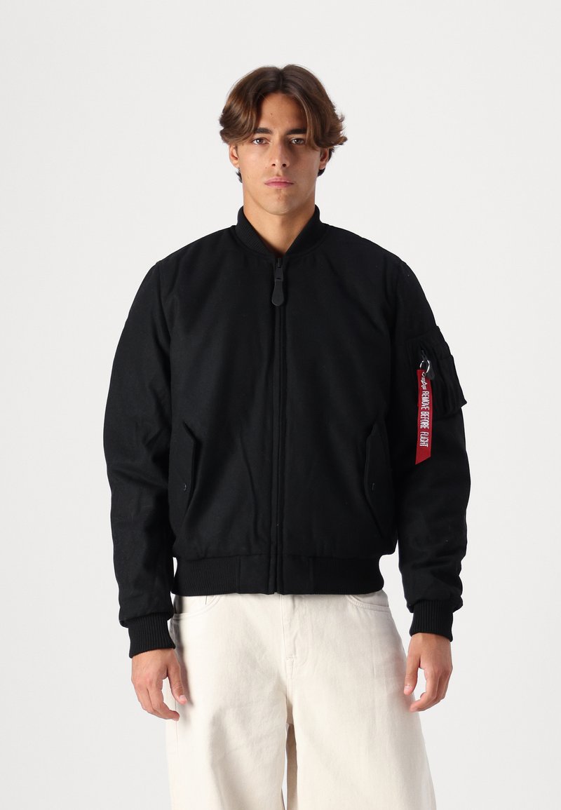 Schwarze Bomberjacke mit Reißverschluss, gerippten Bündchen und Saum, seitlichen Taschen und einem roten Etikett auf dem linken Ärmel. Hergestellt aus Bio-Baumwolle.