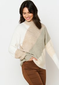 Maglione lavorato a maglia con collo alto, in beige, bianco e grigio chiaro con motivi geometrici e polsini a costine. Abbinato a pantaloni marroni.