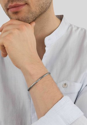 Hombre que lleva una camisa blanca con un cuello pequeño sostiene su barbilla, mostrando una pulsera delgada de cadena de plata en su muñeca.