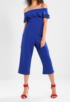 Kvinde iført en royalblå off-shoulder jumpsuit med flæser, vidde bukseben der er korte, og røde høje hæle med anklerem.