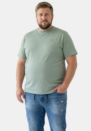 Mann mit Bart, trägt ein hellgrünes T-Shirt und blaue Jeans, steht mit einer Hand in der Tasche vor einem einfarbigen Hintergrund.