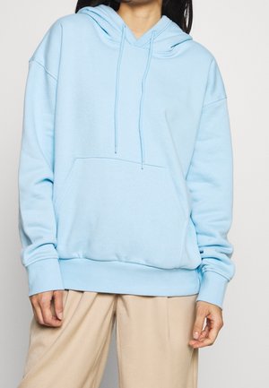 Persoon draagt een lichtblauwe hoodie met voorzak en beige broek, zichtbaar van schouders tot heupen tegen een effen achtergrond.