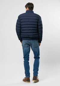 Hombre de pie, vistiendo una chaqueta acolchada azul marino, jeans azules y botas de cuero marrón, de espaldas contra un fondo claro y sencillo.