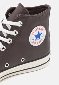 Brown high-top sneaker made of leather with white laces, silver metal eyelets, and a round blue and red logo patch on the side.