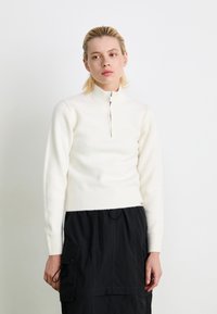 White zip-neck sweater made of soft material, with ribbed cuffs and hem. Worn with a black skirt featuring cargo pockets.