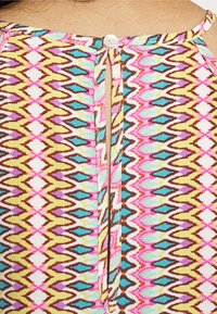 Camisa multicolorida com um padrão geométrico em rosa, azul, castanho e amarelo. Inclui um fecho de botão e uma fenda nas costas.