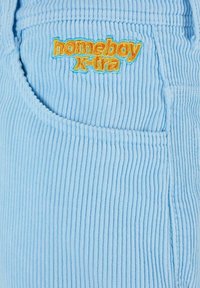 Light blue corduroy fabric with vertical lines, featuring an embroidered "homeboy x-tra" logo in orange and blue on the pocket.