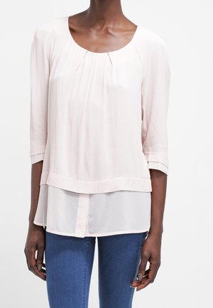 Blusa rosa chiaro con scollatura rotonda, fronte plissettato e maniche a tre quarti. Design a strati con un sottopannello bianco a contrasto.