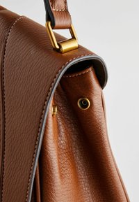Sac à main en cuir marron avec une surface texturée, des accessoires en métal doré et des bordures grises. Il dispose d'une fermeture à pression sécurisée et d'un attachement de sangle robuste.