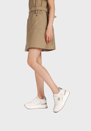 Donna che indossa un vestito beige con cintura e sneakers con plateau bianche con dettagli argentati, in piedi davanti a uno sfondo grigio chiaro uniforme.
