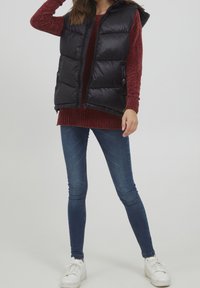 Svart pufferväst med huva, med dragkedjestängning och sidofickor, bärs över en bourgognytt stickad tröja och blå skinny jeans.