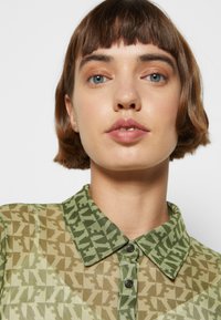 Camisa colarinho com um padrão geométrico verde e bege. Material transparente, botões pretos e design de colarinho contrastante.