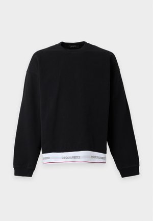 Homme debout portant un sweat-shirt noir, un pantalon beige à jambes larges, des chaussures noires et une ceinture blanche de marque visible au-dessus du pantalon.