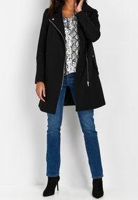 Černý kabát se zapínáním na zip, bočními kapsami a přizpůsobeným střihem. Nosený přes lehký vzorovaný top a doplněný modrými džínami a černými podpatky.