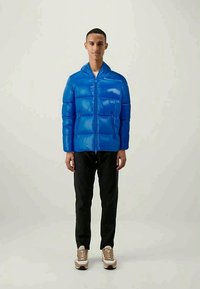 Μπλε φουσκωτό μπουφάν με γυαλιστερή επιφάνεια, φερμουάρ μπροστά, κουκούλα και δύο πλαϊνές τσέπες, συνδυασμένο με μαύρο παντελόνι και μπεζ αθλητικά παπούτσια.