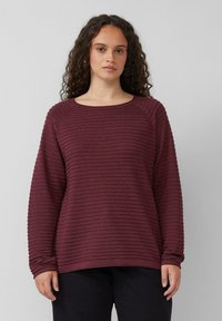Frau mit lockigem Haar, die einen weinroten, gerippten Langarm-Pullover und schwarze Hosen trägt, steht vor einem schlichten, hellgrauen Hintergrund.