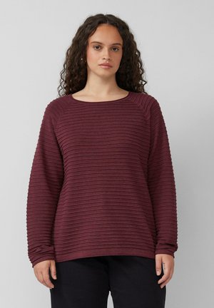 Femme aux cheveux bouclés portant un pull à manches longues côtelé bordeaux et un pantalon noir, se tenant devant un fond gris clair uni.