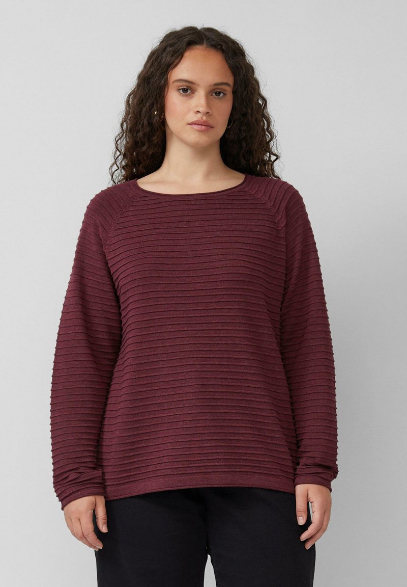 Frau mit lockigem Haar, die einen weinroten, gerippten Langarm-Pullover und schwarze Hosen trägt, steht vor einem schlichten, hellgrauen Hintergrund.