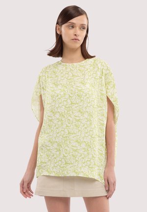 Kobieta o prostych brązowych włosach, ubrana w luźną, jasnzieloną bluzkę w kwiaty z rozciętymi rękawami oraz beżową spódnicę, patrząca na bok.