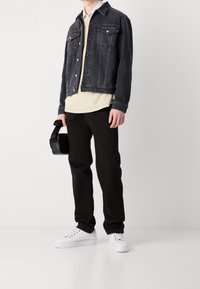 Svart denimjacka över en beige tröja, kombinerad med svarta byxor och vita sneakers. Håller en liten svart väska.