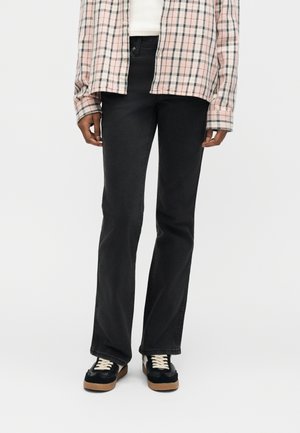 Personne portant un jean noir à jambes droites, des baskets noires et blanches, et une chemise à carreaux rose et blanche par-dessus un haut blanc.