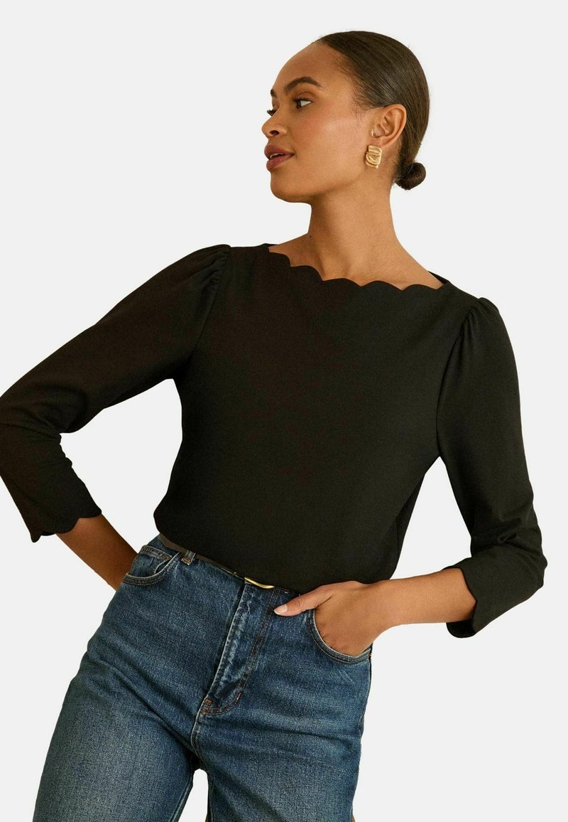 Fekete blúz, hullámos dekoltárral, háromnegyedes ujjal és strukturált anyaggal, közepes mosású, magas derekú farmernadrággal párosítva.